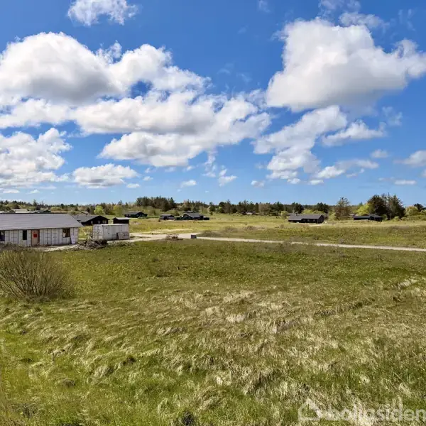 En simpel hytte står stille i et åbent, grønt landskab med græs og buske under en blå himmel med spredte, hvide skyer. Der er ingen tekst på billedet.