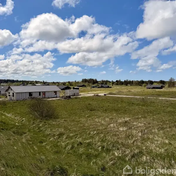 Hus under opførelse står stille på en åben mark med blå himmel, spredte skyer og omkringliggende bygninger i baggrunden. Græsset er grønt og frodigt.