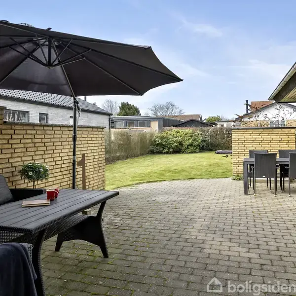 Terrasse med sort parasol og grill. Havebord med stole på stenbelagt område. I baggrunden ses græsplæne og hæk i et boligområde. Ingen tekst fundet i billedet.