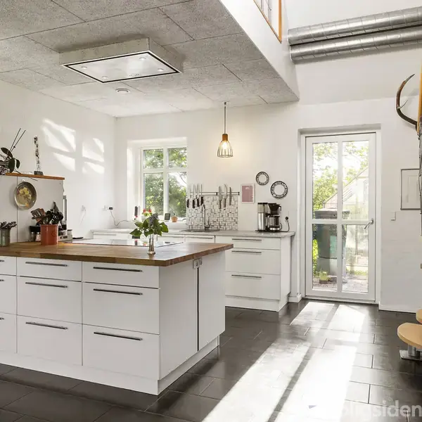 Køkken med hvidt køkkenbord og vask i midten har lys hængende fra loftet. Trappe til højre fører til ovenpå niveau. Vinduer giver kig til have udenfor.