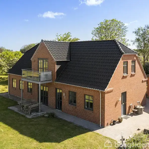 Et rødt murstenshus med sort tegltag har en balkon og store vinduer. Hagen er græsbeklædt med spredte buske og træer omkring. Skyfri himmel og sollys danner en lys atmosfære.
