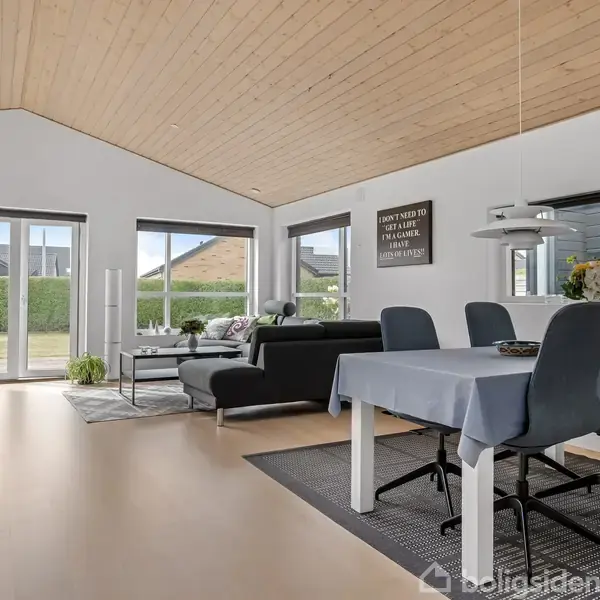 En moderne stue med en sort sofa, placeret ved store vinduer med udsigt til en have. En spisestue med fire stole og bordlampe er til højre. Væggens tekst: "I don't need to flirt, I will seduce you with my awkwardness".