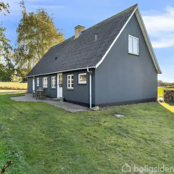 Et sort hus med skråt tag står på en grøn græsplæne. Huset har hvide vinduer og døre, omgivet af træer og en stengærde i en landlig setting.