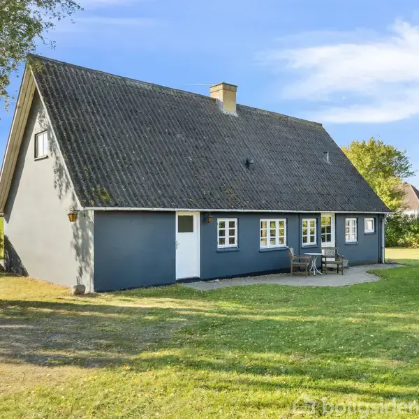 Et gråt hus med skråt tag står stille i en frodig have med træer og græsplæne. En lille terrasse har havemøbler ved siden af døren.