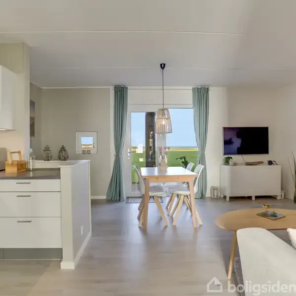 Et moderne opholdsrum med åbent køkken og stue. Køkkenet har hvide skabe og en mikrobølgeovn. Stuen har et sofabord, fladskærms-tv og spisebord nær en glasdør med udsigt til grønt område.