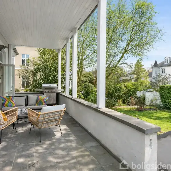 En overdækket terrasse har to flettede stole og et sofamøbel med farverige puder. Terrassen vender ud mod en grøn have med hække, træer og nabohuse i baggrunden.