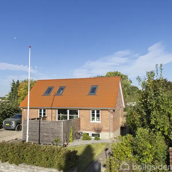 Et rødt murstenshus med en orange tag, placeret i en grøn have. En bil holder parkeret ved siden af huset. Flagstang i forhaven under en klar, blå himmel.