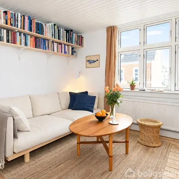 En hvid sofa står i en lys stue ved et træbord med appelsiner og vase med blomster; hylde med bøger hænger over, og vinduer med gardiner slipper dagslys ind.