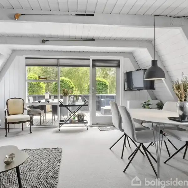 Stue med skråvægge har blå sofa og sofabord til venstre. Hvidt spisebord med stole til højre. Glasdør åbner til udendørs terrasse med grønne planter i baggrunden.