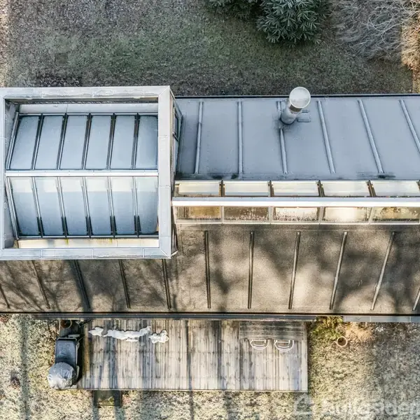 Et rektangulært hus med fladt tag og ovenlys er set fra oven. Ved siden af huset er en terrasse med to personer og en grill. Omgivet af græs og træer.