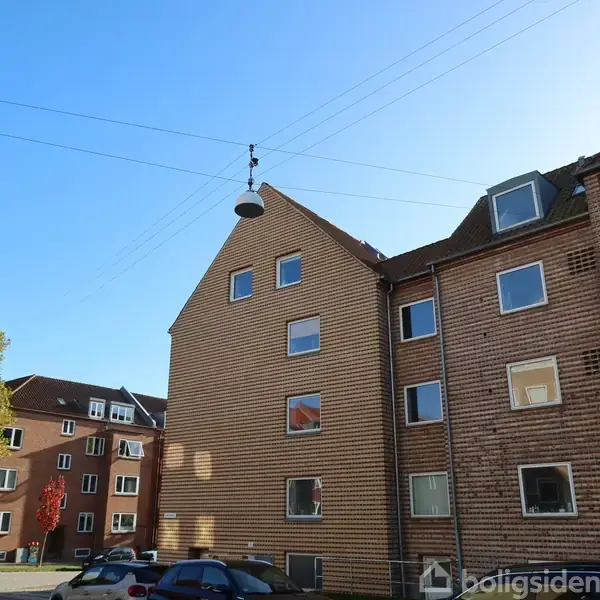 En række af murstensbygninger står under en klar blå himmel. Bygningerne har flere vinduer. Der er biler parkeret på gaden, og træer med efterårsfarvede blade ses i baggrunden.