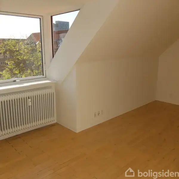Et lyst rum med trægulv og skråvægge, hvor en radiator står under et stort vindue. Udsigt til træer og bygninger udenfor. Ingen tekst til stede.