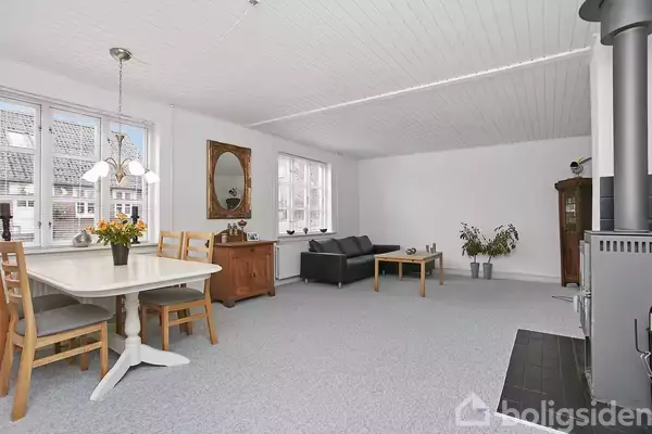 Et lyst rum med spisestue, hvor et bord og stole står ved et vindue. En sofa står i baggrunden. Blomster pryder bordet, og en brændeovn er i hjørnet.