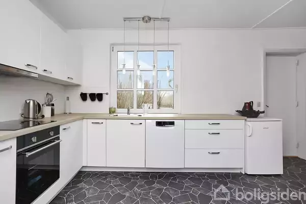 Hvidt køkken med sorte fliser, ovn og opvaskemaskine integreret i skabene. En elkedel og andre småapparater står på bordet, mens et vindue giver naturligt lysindfald.
