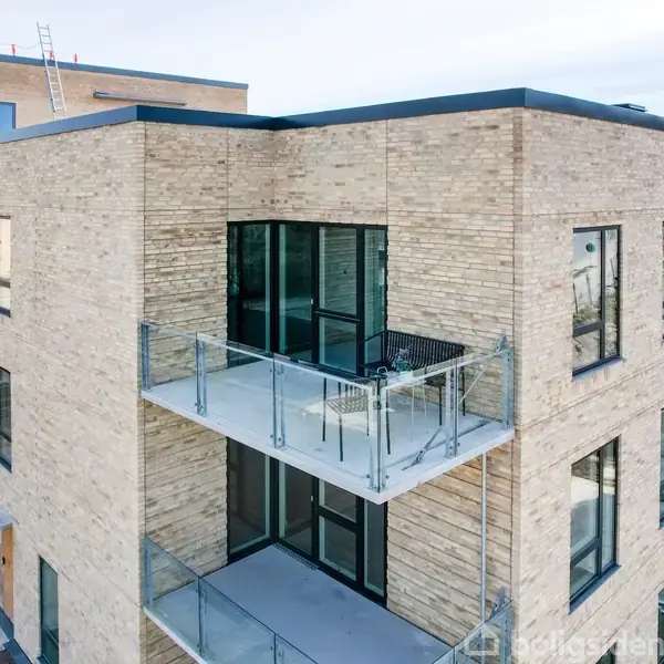 Moderne lejlighedskompleks med lyse mursten og store vinduer; altaner med glasrækværk; beliggende i et urbant område uden synlige tegn på beboelse eller vegetation.