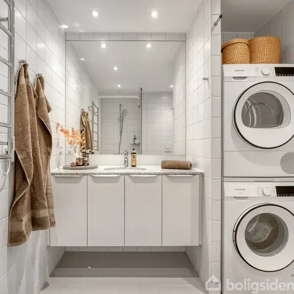 Vaskesøjle med vaskemaskine og tørretumbler står i et lyst badeværelse. Håndklæder hænger på væggen. Indbyggede skabe under spejlet. Kurve er placeret ovenpå maskinerne.