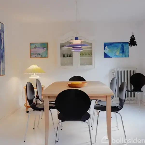 Et spisebord med sorte stole står midt i et lyst rum. Væggene er dekoreret med farverige malerier, og en lampe hænger over bordet. En radiator ses i hjørnet.