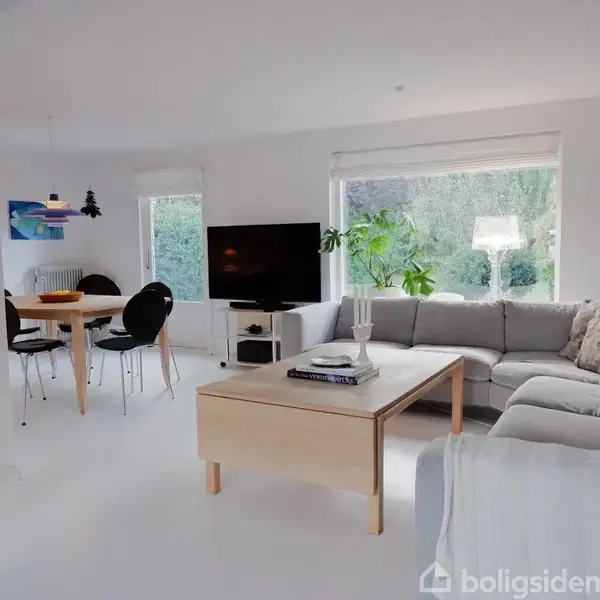 En lys stue med en grå hjørnesofa omkring et træbord. Et TV står ved siden af en stor plante. Et spisebord og stole er i baggrunden.