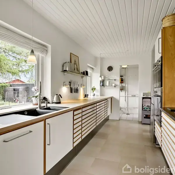 Et moderne køkken med hvide skabe og trædetaljer. En vask, køkkenredskaber og potteplanter står på bordpladen. Store vinduer giver udsyn til en have med grønne træer.