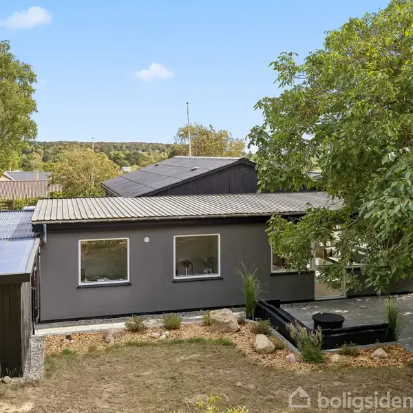 Et gråt hus med fladt tag og fire vinduer står omgivet af træer og en græsplæne i et landligt område under en blå himmel.