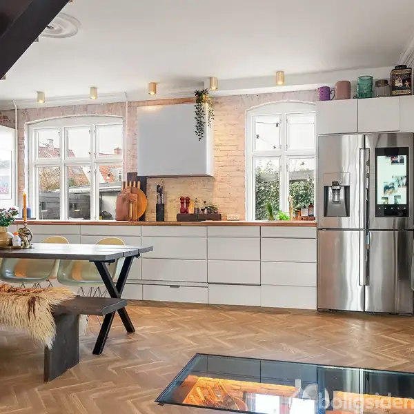 Et moderne køkken med hvidt interiør og stål-køleskab. Bordet har klare stole og en bænk med skind. Trægulv og stor glasplade ved siden af spiseområdet.