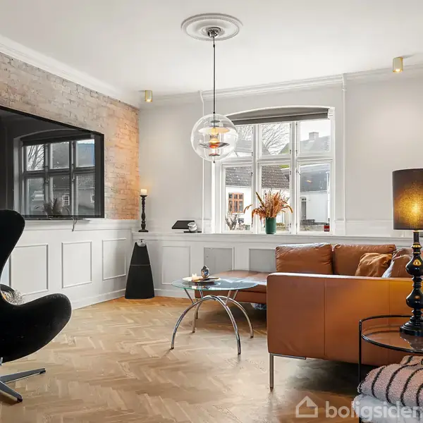 En hyggelig gårdhave med en blå sofa og et boblebad er placeret på en brolagt terrasse. Lyskæder hænger over området, og planter skaber en grøn atmosfære.