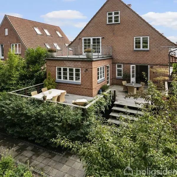 Rødt murstenshus med en stor terrasse, hvor der står havemøbler. Huset er omgivet af grøn beplantning og haveområder, med to naboer i baggrunden under en blå, skyfri himmel.