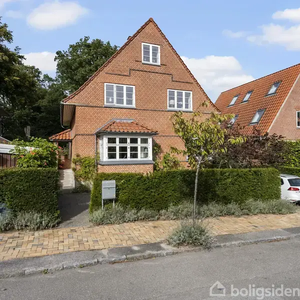 Et rødt murstenshus står stille på en fortovskant omkranset af en lav hæk. Omkring huset ses en brolagt vej, en parkeret bil og nabohuse med røde tage.