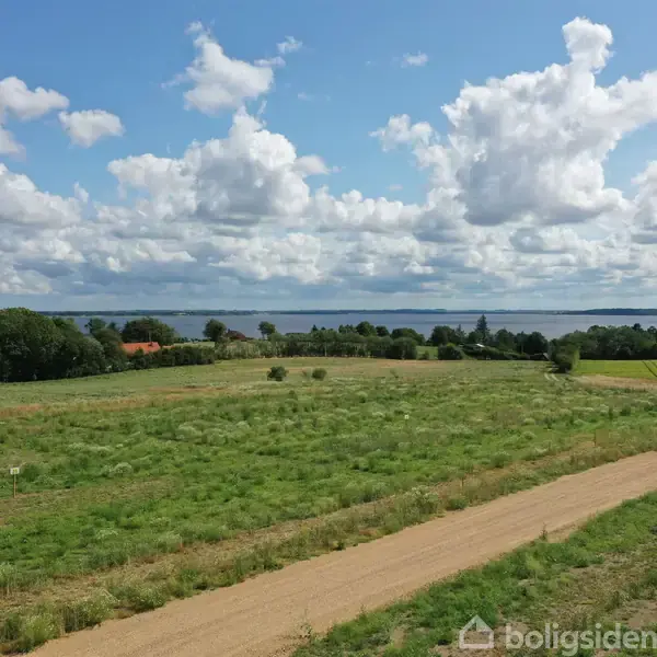 En grøn eng strækker sig mod horisonten med en smal grussti i forgrunden. Træer og en sø danner baggrunden under en let overskyet himmel.