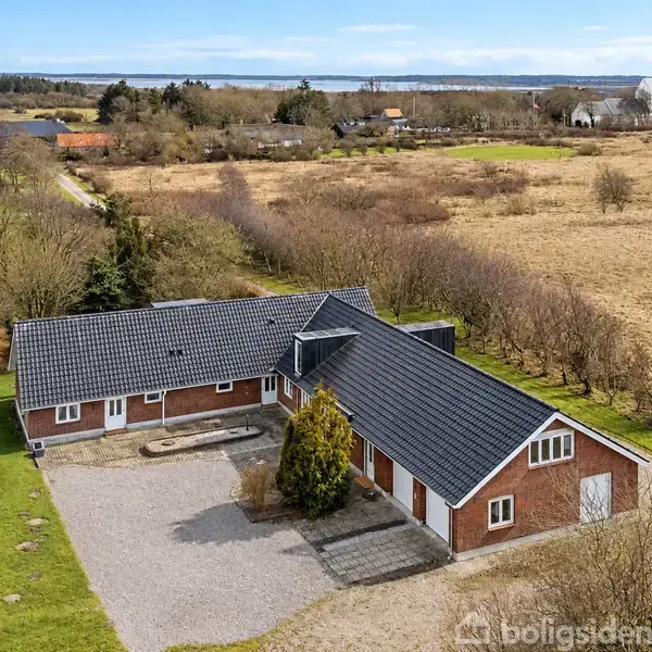 Et rødt murstenshus med antracitgråt tag står på en grusbelagt gårdsplads. Huset er omgivet af åbne marker og en fjern kirke i baggrunden.