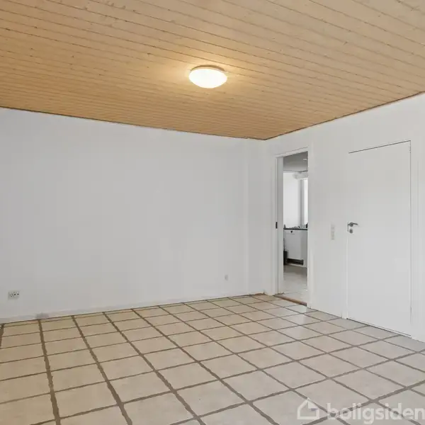 Et rum med lyse fliser på gulvet. Et rund loftlampe lyser hvidmalede vægge og et træloft. En åben dør fører til et tilstødende rum.