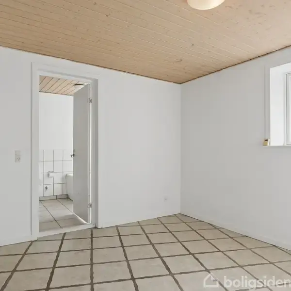 Et tomt rum med flisegulv og træloft, viser en åben døråbning til et flisebelagt badeværelse. Et vindue på væggen giver naturligt lys.