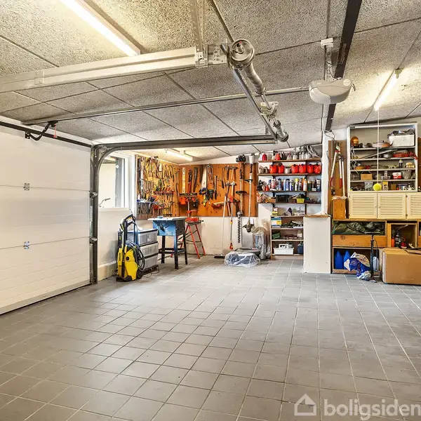Garage med åbent rum, værktøjsophæng på væggen, opbevaringshylder fyldt med forskellige redskaber og materialer. Stort vindue til venstre og flisegulv. Arbejdsbænk placeret under værktøjet.