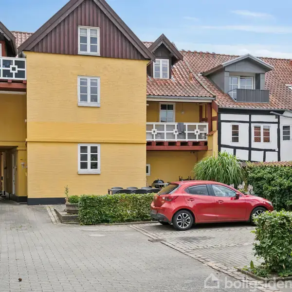 Gul bygning med hvide vinduer står stille, omgivet af en gårdsplads med brosten og en rød bil parkeret langs en lav hæk, under en blå himmel.