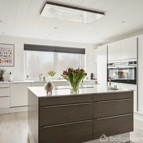 Køkkenbord med tulipaner står i moderne køkken med hvide skabe, indbyggede ovne og murstensvæg. Sollys strømmer ind fra vinduet over håndvasken, og en farvet plakat hænger på væggen.