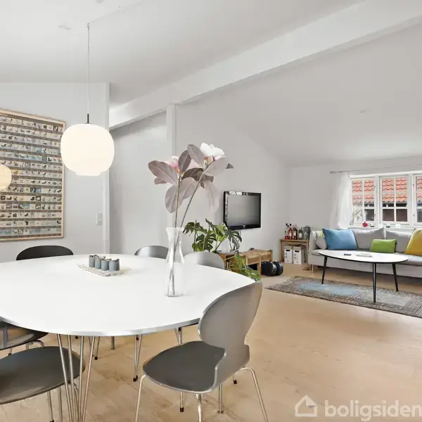 Rundt spisebord med stole står i et lyst rum; en vase med blomster i midten. Sofagruppe ved vinduet med farverige puder. Moderne kunst hænger på væggen.