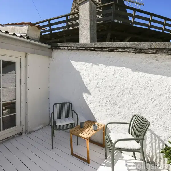 En terrasse med to stole og et lille bord, stående på en lys træplatform. Omkranset af en hvid mur, med en bygning i baggrunden under en klar blå himmel.