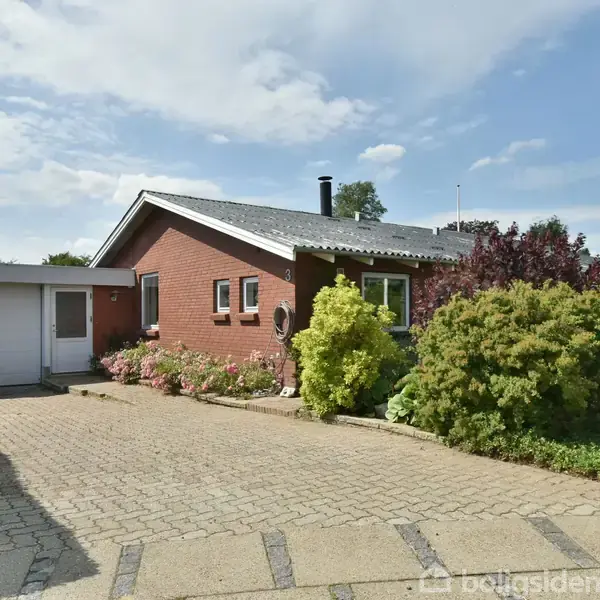 Rødt murstenshus med fladt tag og tilhørende carport, omgivet af velplejede buske og græsplæne. Foran huset er en bred flisebelagt indkørsel. Skyfri himmel i baggrunden.