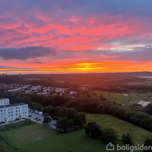 Solnedgang farver himlen i pink og orange over en by med hvidgrå bygninger, grønne træer og marker. Skyernes formationer giver billedet et dramatisk udtryk.