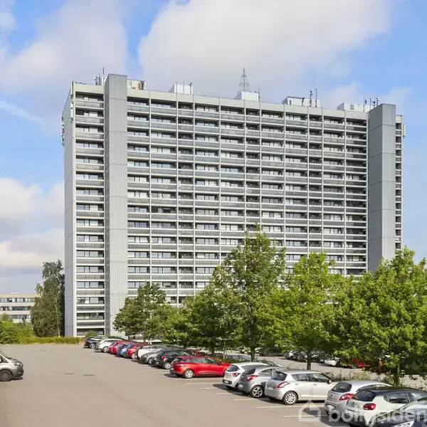 En høj boligblok med mange altaner står stille omgivet af parkerede biler og grønne træer på en parkeringsplads. Skyer ses på himlen i baggrunden.