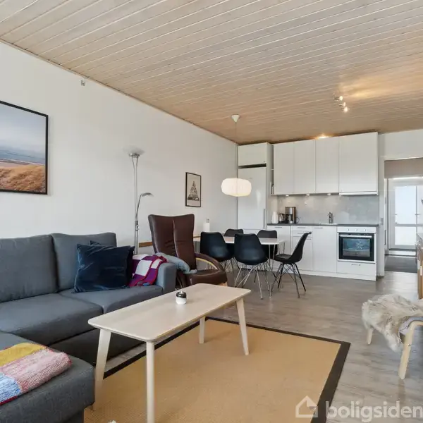 Grå sofa står i en stue med et kaffebord foran. Køkkenområdet med hvidt køkkenskab er i baggrunden, og et spisebord med sorte stole er placeret ved siden.