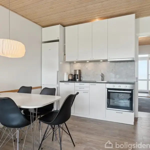 Et hvidt køkken med indbygget ovn og køleskab. Ved siden af står et ovalt spisebord med sorte stole. En lampe hænger over bordet. Billedet er fra Boligsiden.