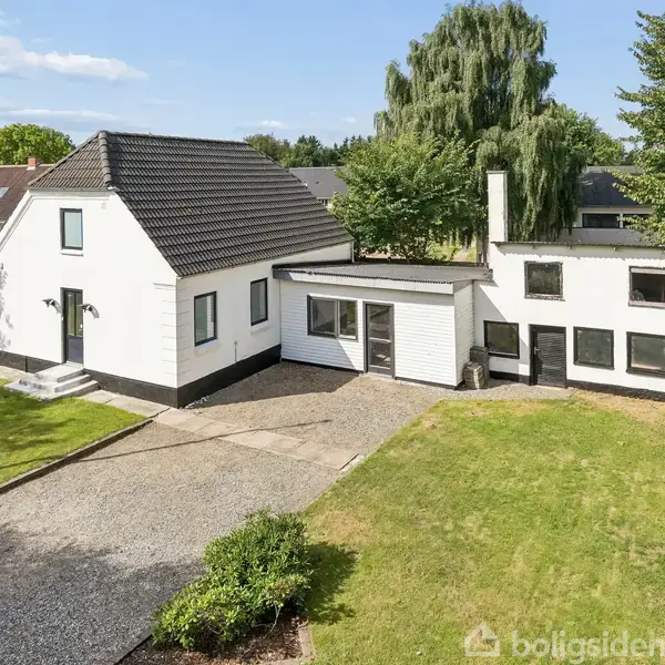 Hvidt hus med sort tag står stille i grøn have med grusindkørsel. Omgivet af træer og græsplæne, under blå himmel med få skyer.