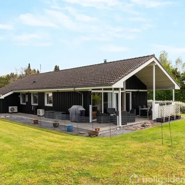 Et sort træhus står på en stor græsplæne med hække i baggrunden. Huset har en overdækket terrasse med siddepladser, omgivet af grønne træer og buske under en blå himmel.