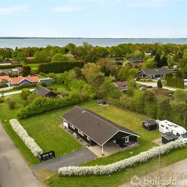 En etplanshus omgivet af græsplæne og buske står ved en vej. En autocamper er parkeret ved indkørslen, og omgivelserne er grønne med udsigt til en stor sø i horisonten.