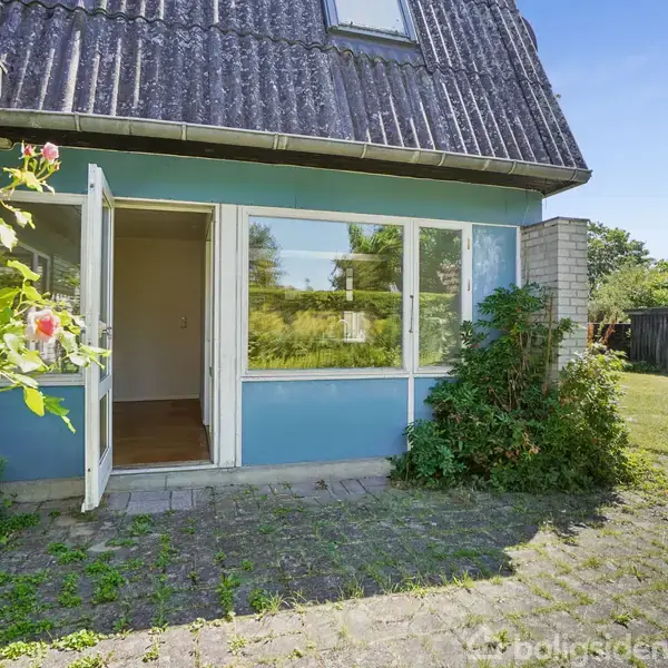 Hus med blå facade og åbne døre. En terrasse med grå fliser foran huset. Omgivet af have med græsplæne og buske.