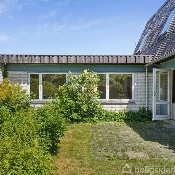 Hus i lys stenen med mange vinduer; en havedør er åben. Haven har grønne buske og græs. Klar blå himmel i baggrunden.