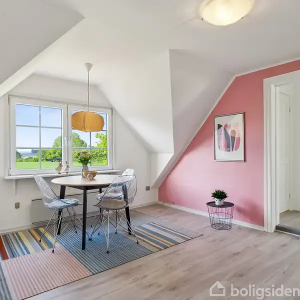 Rundt bord med fire stole står ved vindue. Bordet har en pendellampe over. Væggen er malet lyserød, og gulvet har et stribet tæppe. Et maleri hænger på væggen.