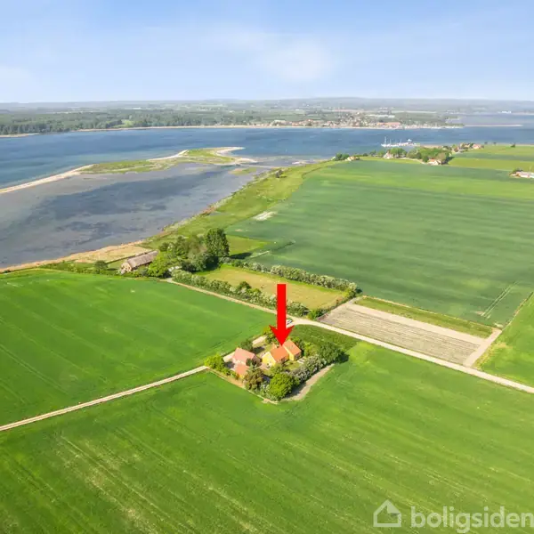 Hus markeret med en rød pil; omgivet af grønne marker. I baggrunden ses kystlinje og hav. Klar blå himmel og landlig atmosfære.