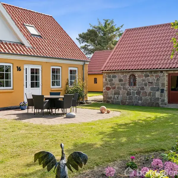 Et gult hus med nummer 16 har en terrasse med bord og stole. En stenbygning med rødt tag står i baggrunden. Haven har blomster og en fuglefigur.